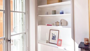 Um quarto com parede branca, janela com moldura branca, uma prateleira com diversos objetos e porta com moldura branca.