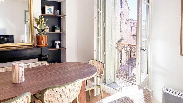Quarto moderno com mesa de jantar, cadeiras, um espelho grande e vista para um edifício através de uma porta aberta.