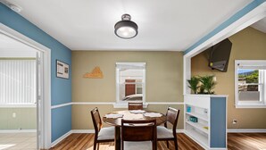 A dining area with a round table, four chairs, a ceiling light, a window with blinds, a wall-mounted TV, and a plant. - Family Beach House · Sleeps 6 · 2 Blks · 6 Parking (Virginia Beach)