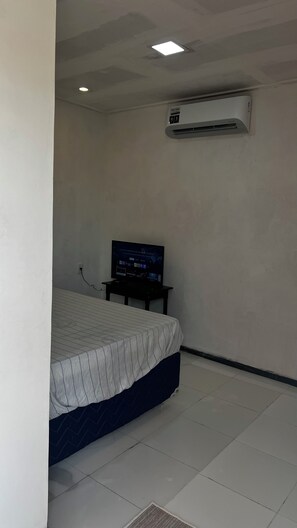 A bedroom with a bed, a television on a stand, an air conditioning unit on the wall, and a tiled floor.