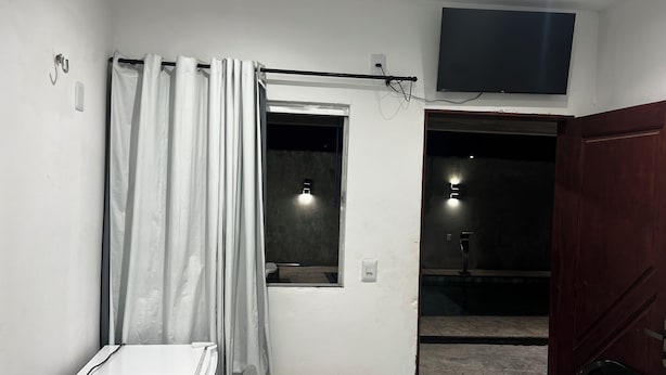Ein Zimmer mit weißer Wand, einem Fenster mit Vorhang, einem flachbildfernseher über dem Fenster und einer Holztür.