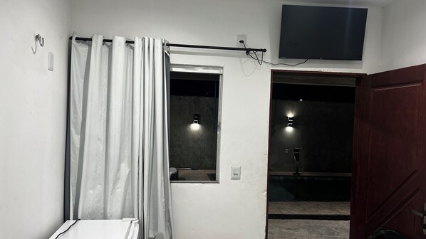 A room with a white wall, a window with a curtain, a flat-screen TV mounted above the window, and a wooden door.