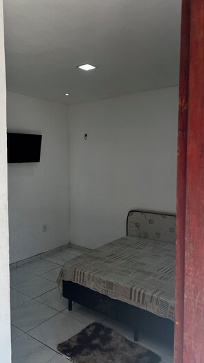 A small, sparsely furnished room with a bed, a flat-screen TV mounted on the wall, and a tiled floor.