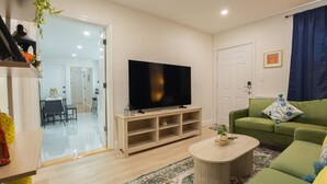 Een woonkamer met een flatscreen televisie, een groene bank, een salontafel en een eetgedeelte zichtbaar door een glazen deur.