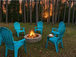 Ein Lagerfeuer mit vier türkisfarbenen Adirondack-Stühlen, die darum herum in einer Wiese platziert sind, mit Wald im Hintergrund.