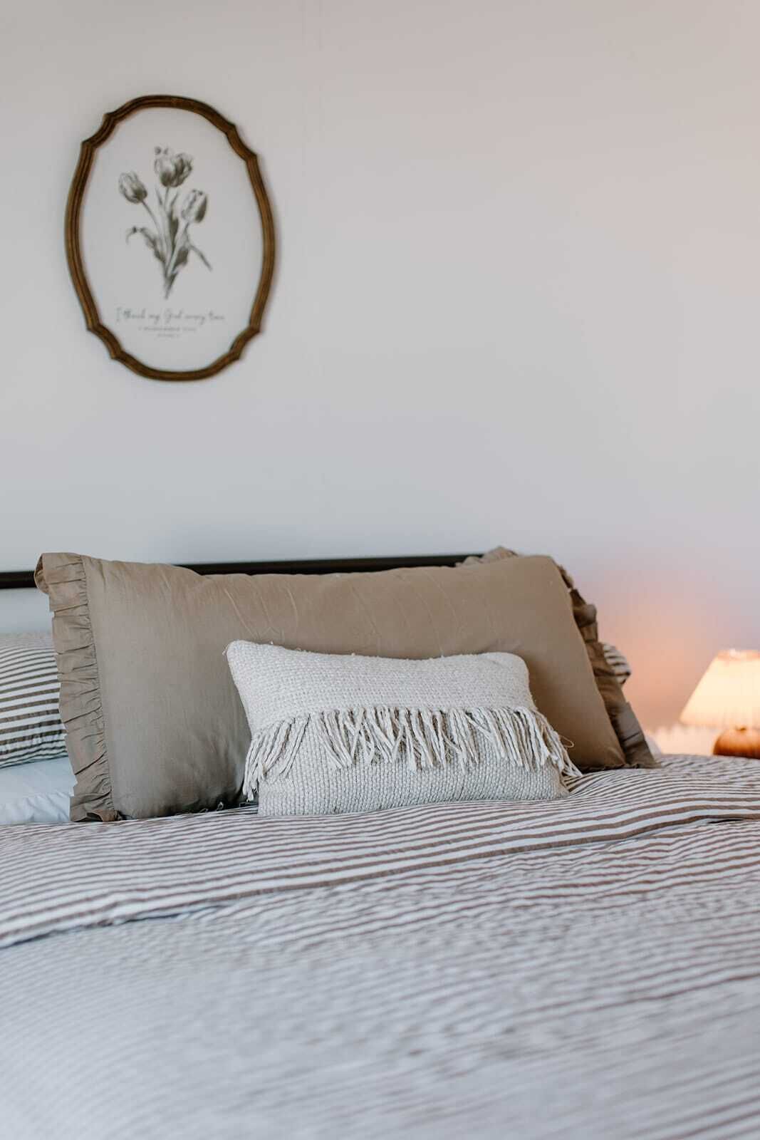 A bed with striped bedding, a beige pillow, and a white pillow with fringe.