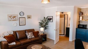 A living room with a brown leather sofa, a round coffee table, and a small side table with a lamp. There is a door leading to a bathroom, and a kitchen area with blue cabinets and a microwave.