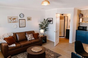 Una sala de estar con un sofá de cuero marrón, una mesa de centro redonda y una mesita auxiliar con una lámpara. Hay una puerta que da a un baño, y una zona de cocina con armarios azules y un microondas.