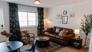 A living room with a brown leather sofa, a round coffee table, a dining table with chairs, a lamp, and a potted plant.