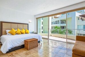 A bedroom with a large bed, a small side table, a wicker basket, and a view of a balcony with buildings and greenery. - Spacious Penthouse with ocean views, pool & balcony (Nuevo Vallarta)