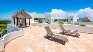 Una terraza en la azotea con sillones de descanso, una estructura con techo de paja y edificios modernos al fondo.