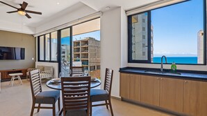 A modern kitchen with a large window, a dining area with chairs, and a ceiling fan. - Modern condo with infinity pool, WiFI, plunge spa & ocean-view balcony (Nuevo Vallarta)