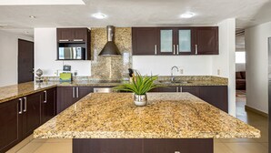 A modern kitchen with granite countertops, dark cabinetry, stainless steel appliances, and a central island with a potted plant. - Luxurious Penthouse with a scenic terrace pool & modern kitchen (Nuevo Vallarta)