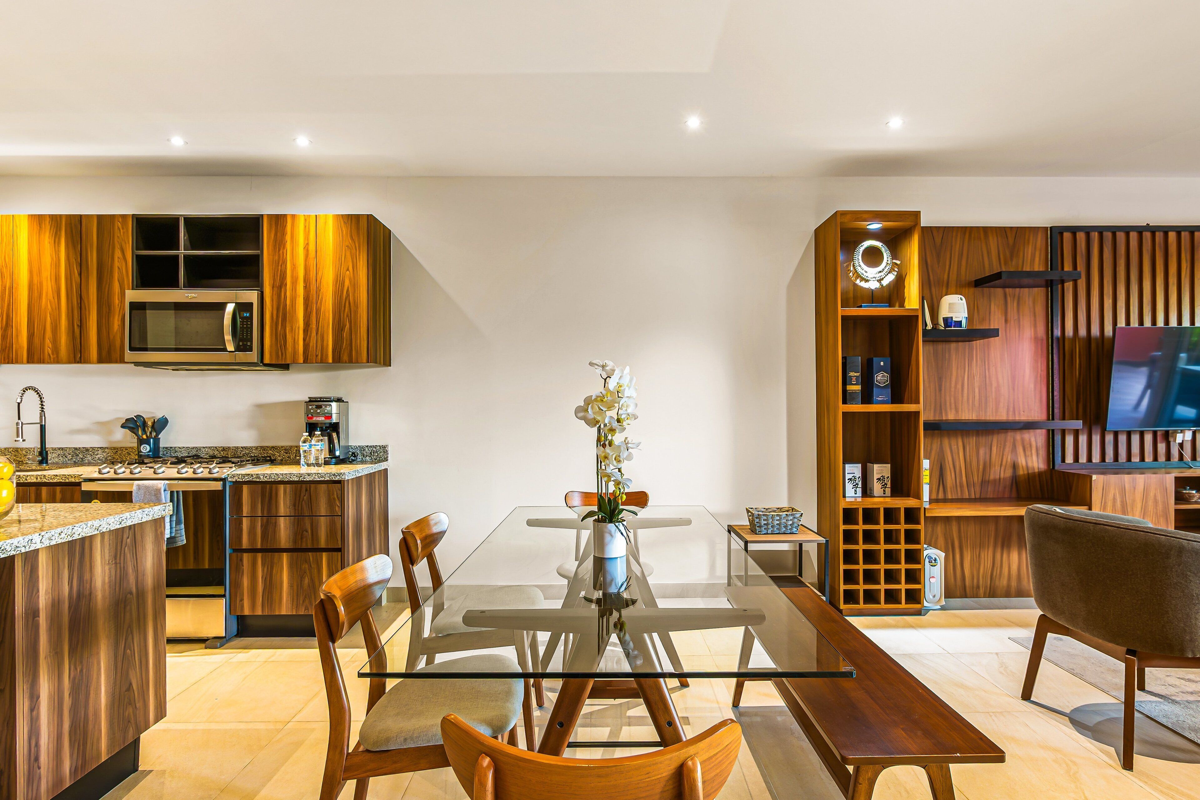 A modern kitchen with a built-in microwave, a dining table with chairs, and a living area with a TV and shelving.