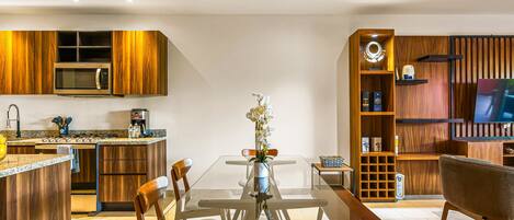 A modern kitchen with a built-in microwave, a dining table with chairs, and a living area with a TV and shelving.