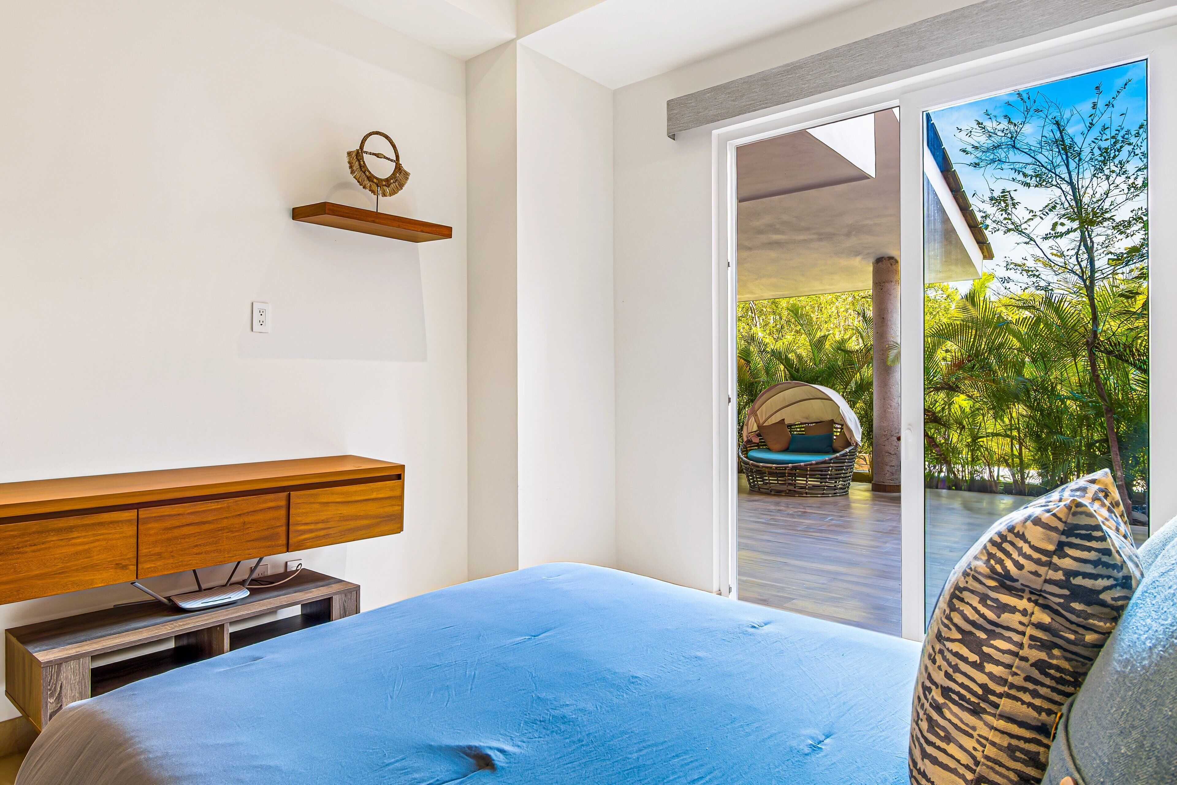 A bedroom with a bed, a wooden nightstand, a wall-mounted shelf, and a sliding glass door leading to an outdoor area with a wicker chair and greenery.