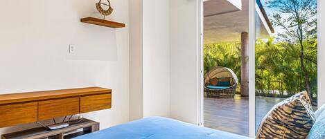 A bedroom with a bed, a wooden nightstand, a wall-mounted shelf, and a sliding glass door leading to an outdoor area with a wicker chair and greenery.