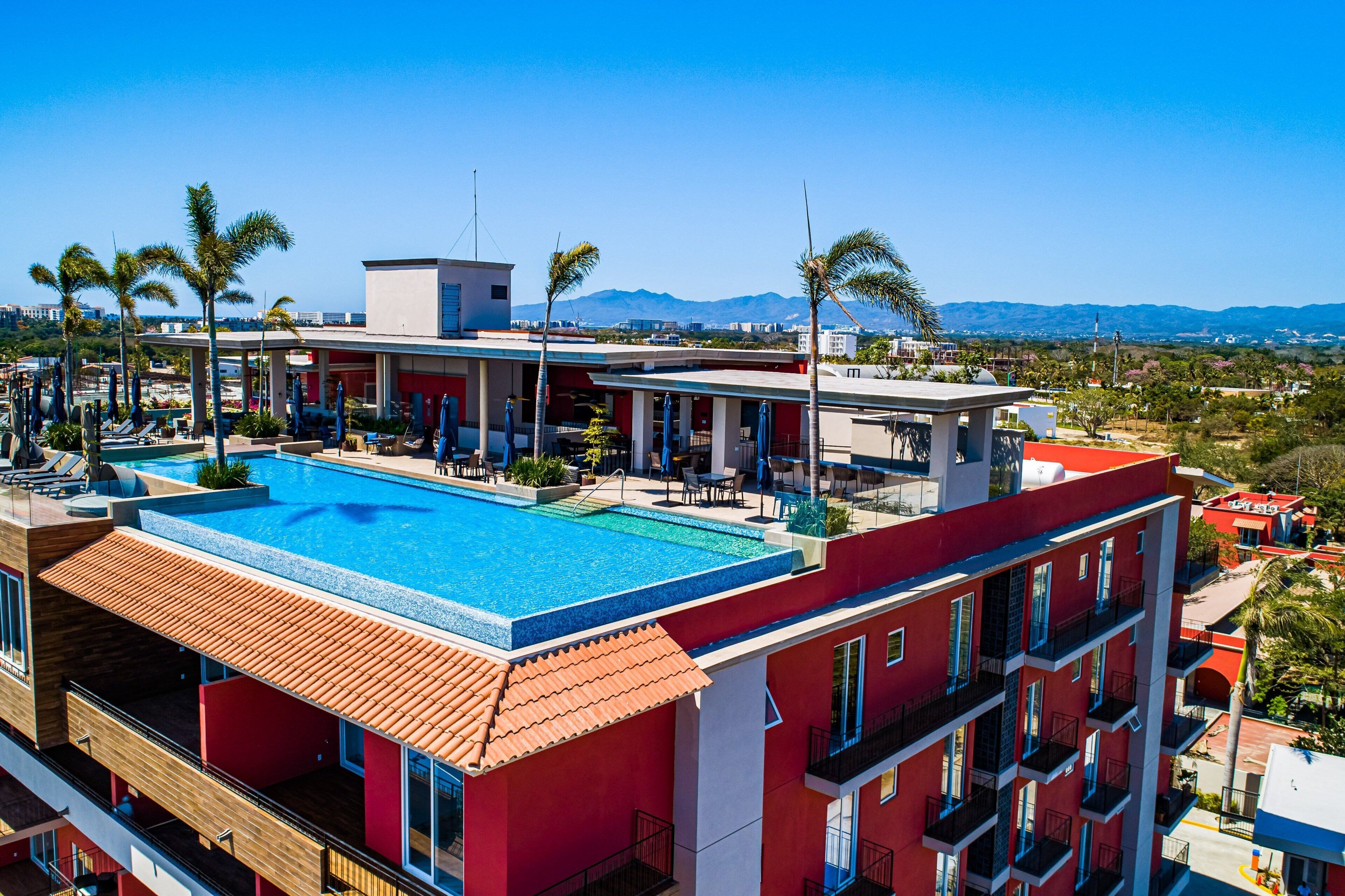 A hotel with a rooftop pool, red and white architecture, and palm trees.