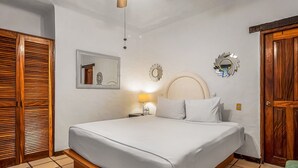 Een slaapkamer met een bed, een houten garderobe, een spiegel, wanddecoratie en een nachtkastje met een lamp.