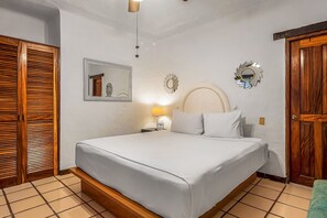 A bedroom with a bed, wooden wardrobe, mirror, wall decor, and a nightstand with a lamp. - Puerto de Luna #622 | 1BR/2BA Puerto Vallarta | Pool, beach & dining (Puerto Vallarta)