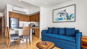 Un salon moderne avec un canapé bleu, des chaises en bois, une table basse et un coin cuisine doté de meubles en bois et d’un réfrigérateur.
