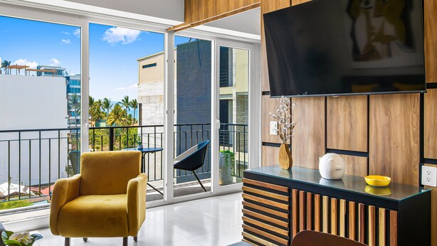 A modern living room with a sofa, a TV, and a balcony with a view of buildings and palm trees. - Viva la vida Life 309 | 1BR/2BA Bucerias, near ocean, pool, balcony, WiFi (Bahia de Banderas)