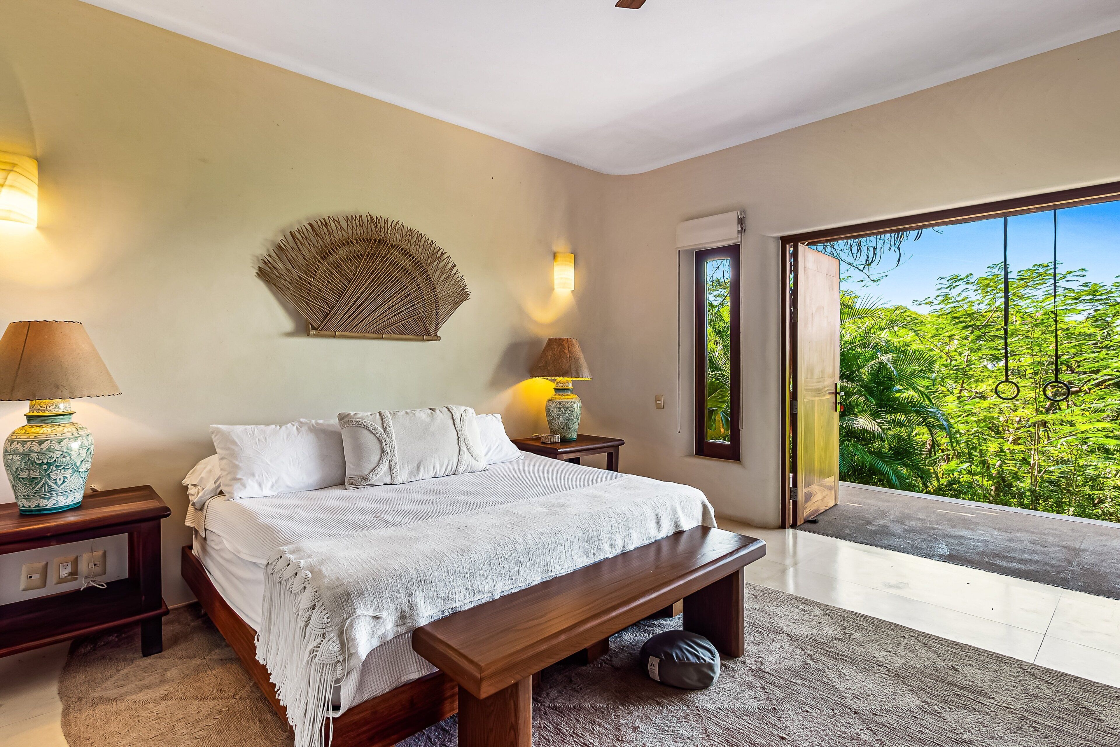 A bedroom with a bed, bedside tables, a bench, and a view of greenery through a large window.