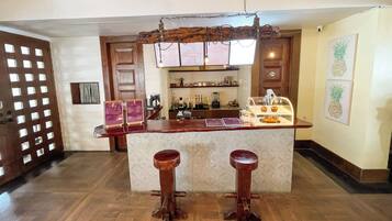 Una zona de bar con mostrador de madera, dos taburetes, un panel con la carta y una obra de arte con forma de piña en la pared.