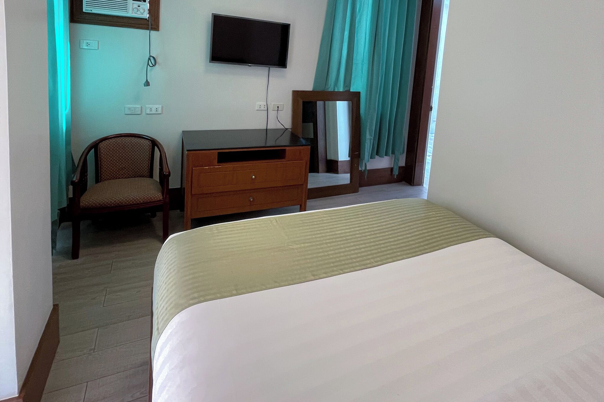 A hotel room with a bed, a wooden dresser, a chair, a television, and a window with curtains.