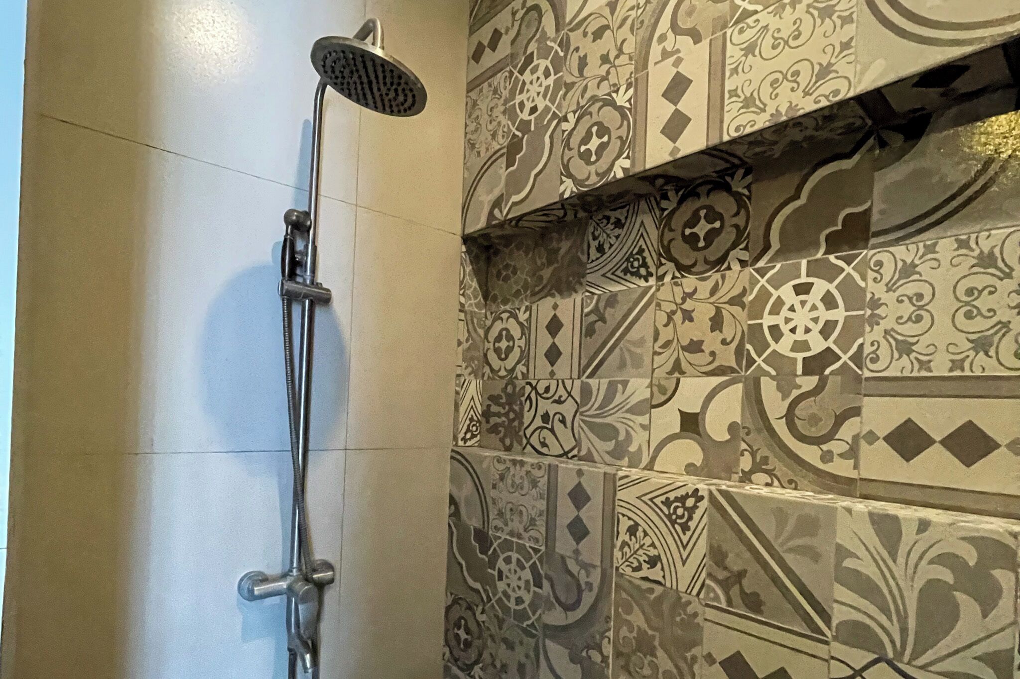 A shower with a handheld showerhead, a wall with patterned tiles, and a plain tiled floor.