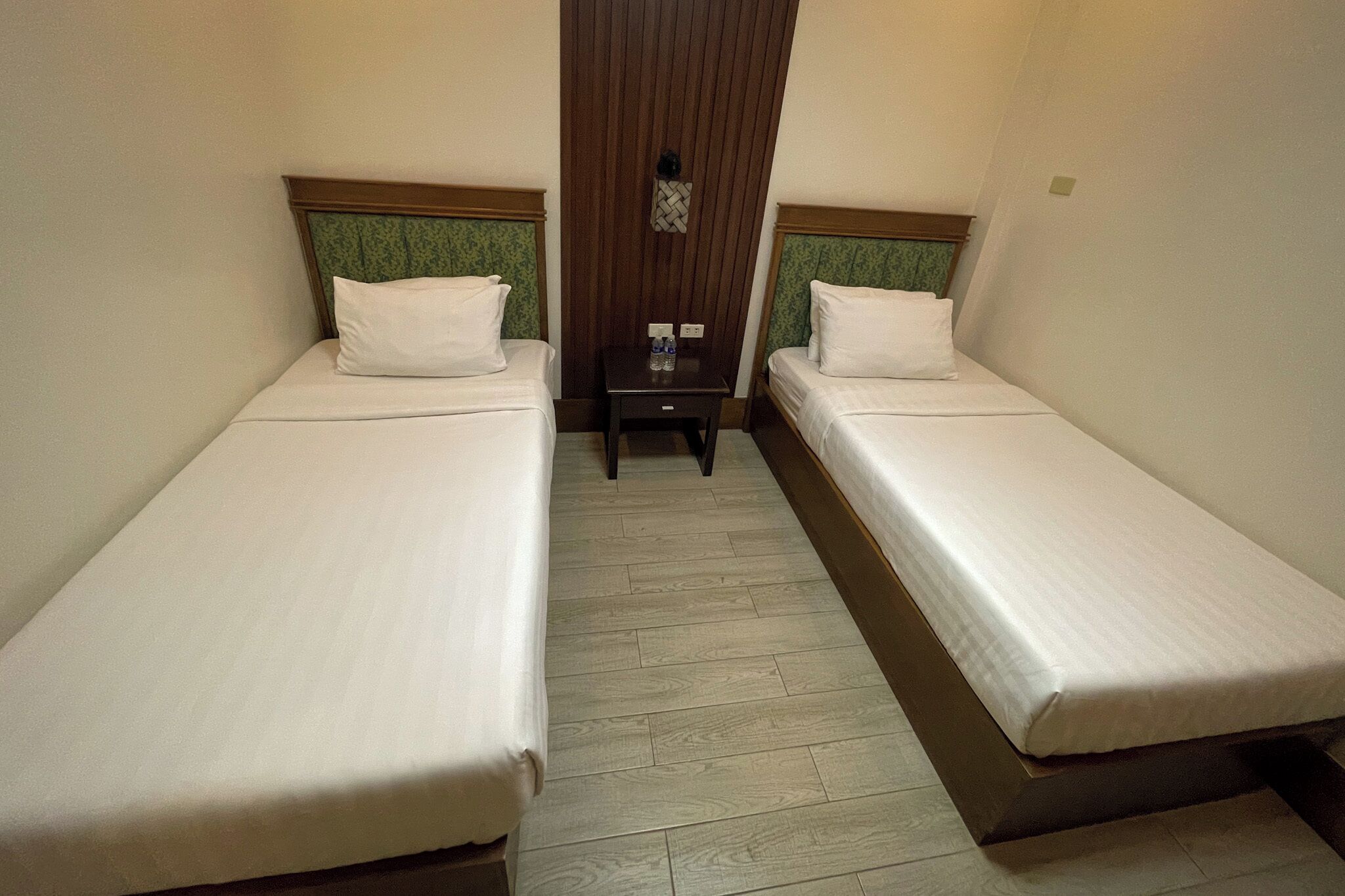 A hotel room with two single beds, a small table with a bottle and a cup, and a wooden floor.