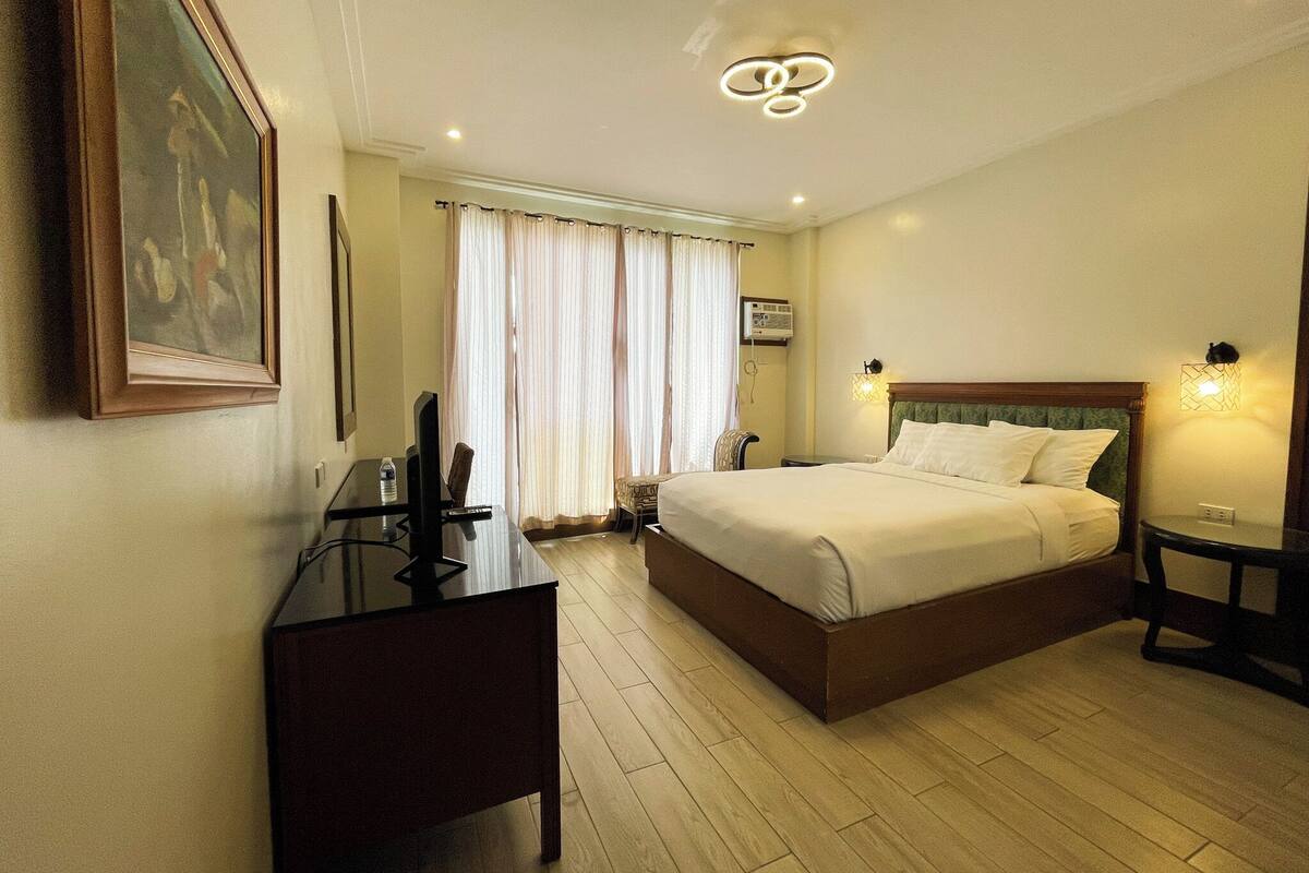 A hotel room with a bed, bedside tables, a desk, a chair, a television, and a painting on the wall.