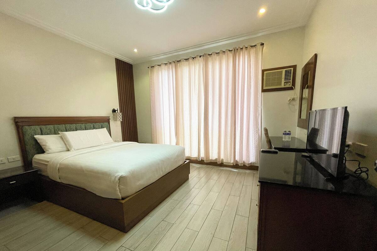 A hotel room with a bed, a desk with a chair, a television, and a window with curtains.