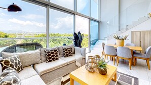 Un salon moderne comprenant un canapé, une table à manger et des chaises, de grandes fenêtres offrant une vue sur la verdure et un paysage urbain lointain.