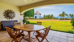 Une terrasse avec une table ronde en bois et des chaises, un ventilateur de plafond, un élément décoratif mural et une vue sur un bâtiment blanc doté d’un toit de chaume.