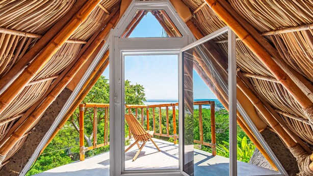 Et trekantet rum med en træstol på en balkon, stråtak og udsigt til grøn natur og havet.