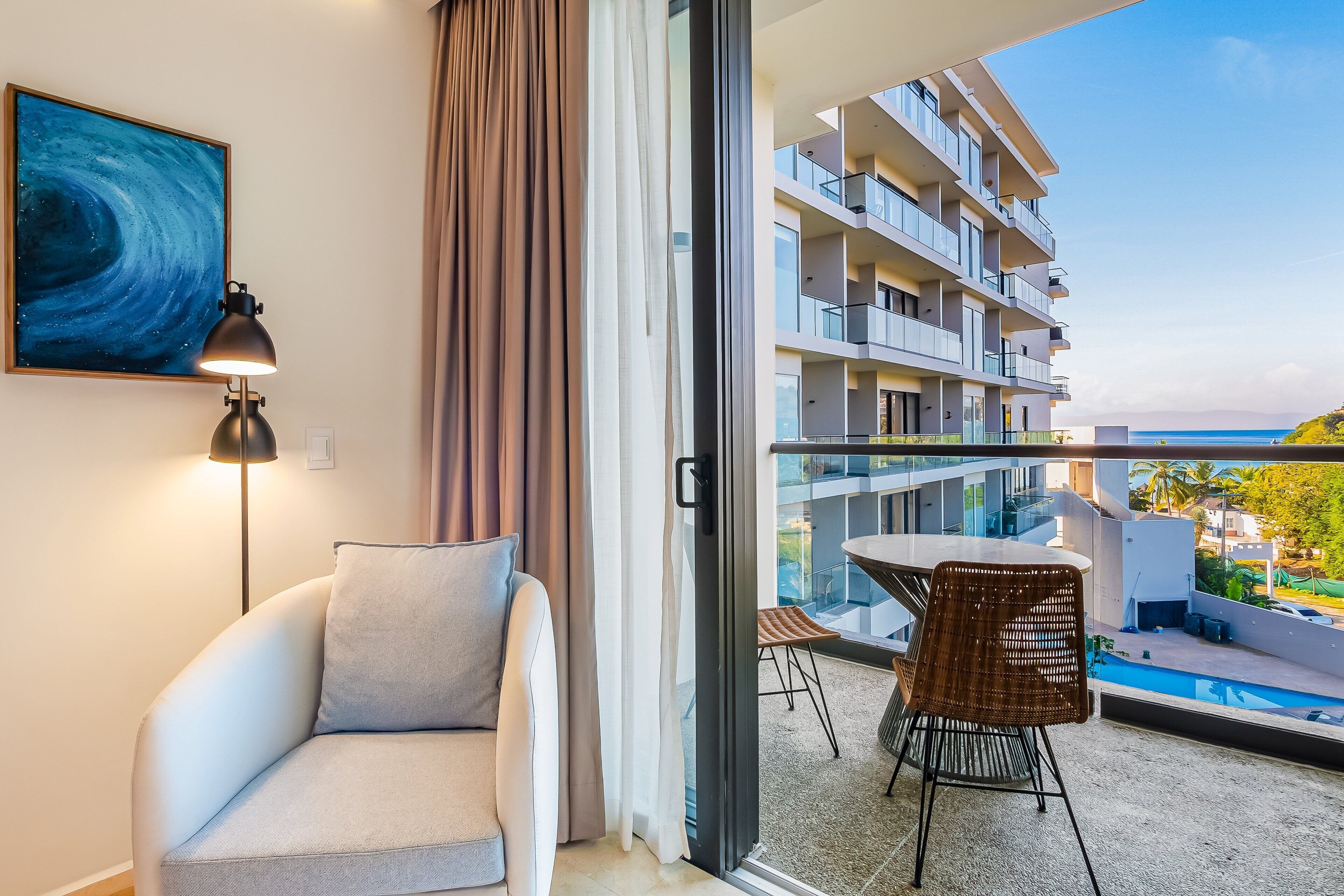 Habitación de hotel moderna con sofá, lámpara, un cuadro, puerta corrediza de vidrio a un balcón con piscina y vistas a otros edificios.