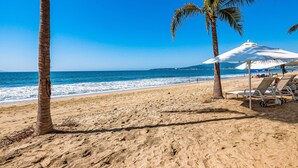 A beach with palm trees, a white umbrella, and lounge chairs. - Ocean-view condo with balcony, central AC, pool, gym, cinema, & bar (Nuevo Vallarta)
