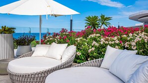 A wicker lounge chair with a white cushion, a white umbrella, and a view of the sea. - Ocean-view condo with balcony, central AC, pool, gym, cinema, & bar (Nuevo Vallarta)
