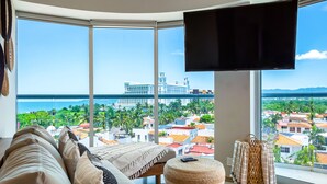 A modern living room with a large window, a flat-screen TV, a sofa, a small ottoman, and a wicker chair. - Ocean-view condo with balcony, central AC, pool, gym, cinema, & bar (Nuevo Vallarta)