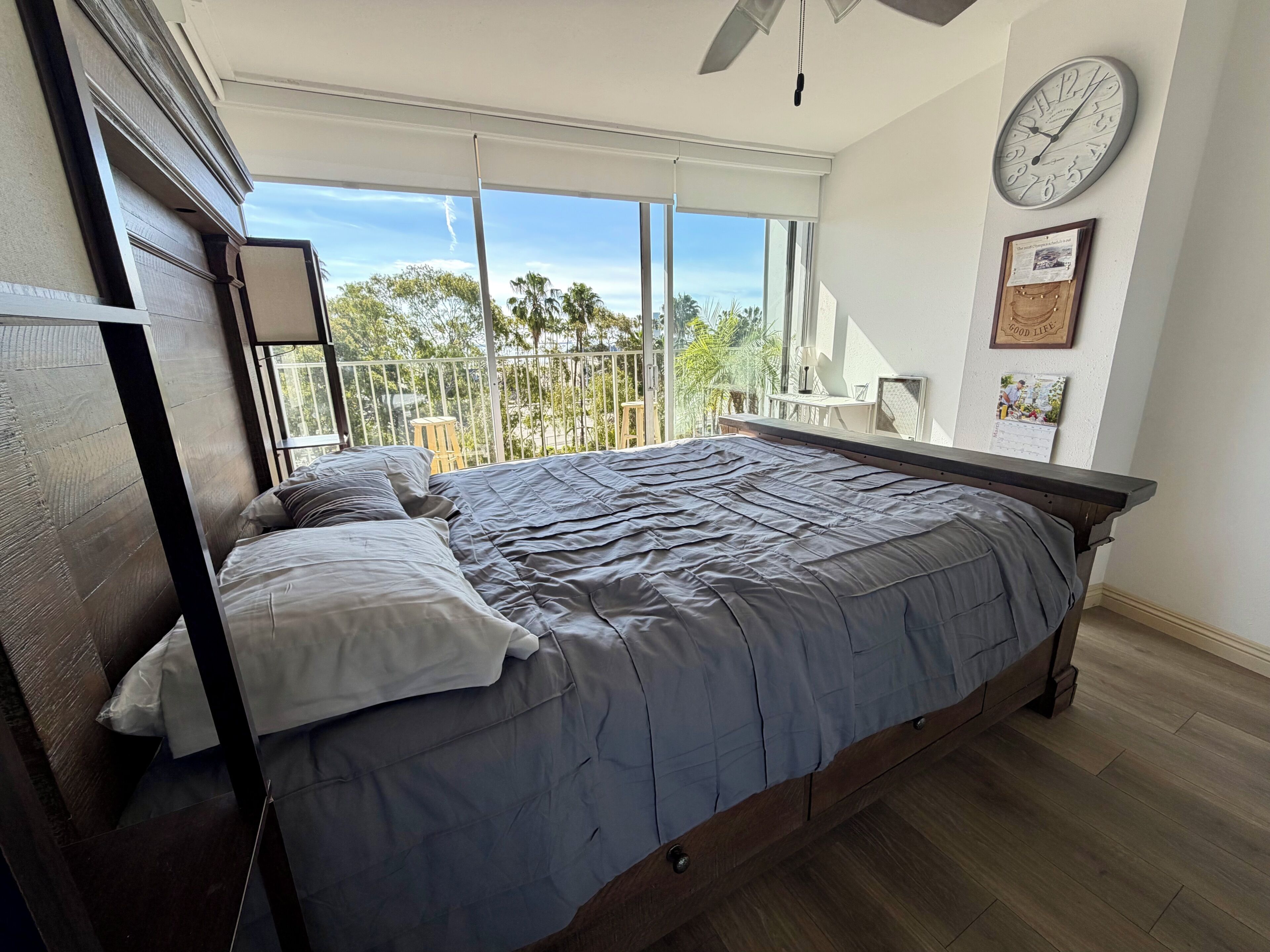Un dormitorio con una cama grande, un ventilador de techo, un reloj y un balcón con vistas a árboles y edificios.