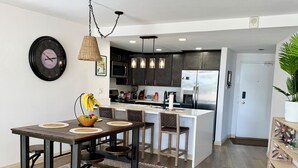 Uma cozinha moderna com armários escuros, uma geladeira de aço inoxidável e uma sala de jantar com mesa e cadeiras de madeira.