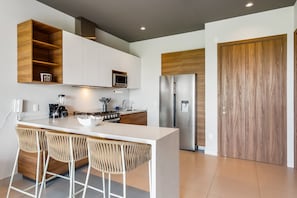 A modern kitchen with a central island, bar stools, a built-in microwave, a stainless steel refrigerator, and wooden cabinetry. - Punto Novo 504 | 1BR Nuevo Vallarta, Near Beach, Pool (Nuevo Vallarta)