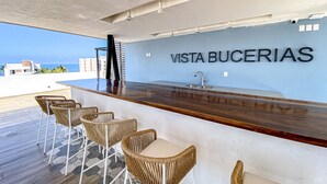 Een barruimte met rieten stoelen en een houten toonbank, een bord met de tekst 'VISTA BUCERIAS' en uitzicht op de oceaan.