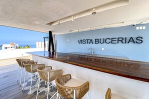 A bar area with wicker chairs and a wooden counter, a sign reading 'VISTA BUCERIAS,' and a view of the ocean. - Ocean-view condo with infinity pool, rooftop lounge, & private balcony (Bucerias)