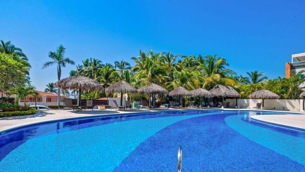 A swimming pool with a clear blue water, surrounded by palm trees and thatched umbrellas. - Luxury Beach Condo | Community Pool, Ocean Views, Walk to Beach (Nuevo Vallarta)