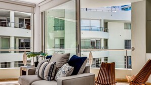 A modern living room with a sofa, cushions, a wooden chair, and a large window overlooking a multi-level building. - Luxury Beach Condo | Community Pool, Ocean Views, Walk to Beach (Nuevo Vallarta)