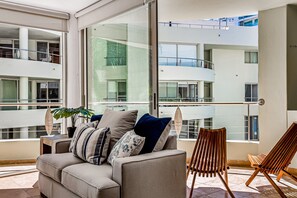 Ein modernes Wohnzimmer mit einer Couch, Kissen, einem Holzsessel und einem großen Fenster mit Blick auf ein mehrgeschossiges Gebäude.