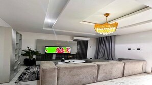 A modern living room with a sectional sofa, a flat-screen TV, a chandelier, and a potted plant. - Supreme Apartments (Ibadan)