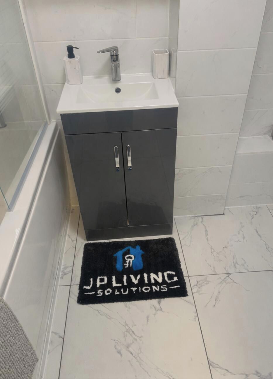 Ett badrum med ett vitt handfat, en spegel och en svart matta med texten ”JALIVING SOLUTIONS”.